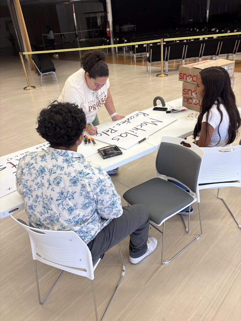 A warm and engaging photo showing Sandra and two youths collaboratively filling out a sign that announces a backpack giveaway for kids. The setting is in Boise, where Love Rain Drops is dediated to supporting local children by providing essential school supplies . This image captures a spirit of community involvment and generosity.